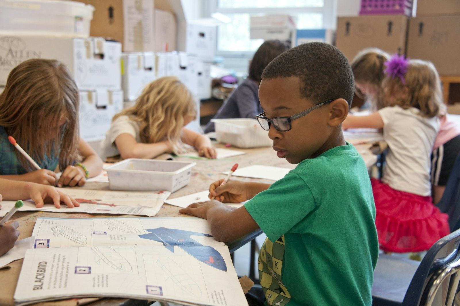 Children work on worksheets at shared tables in a bright classroom.