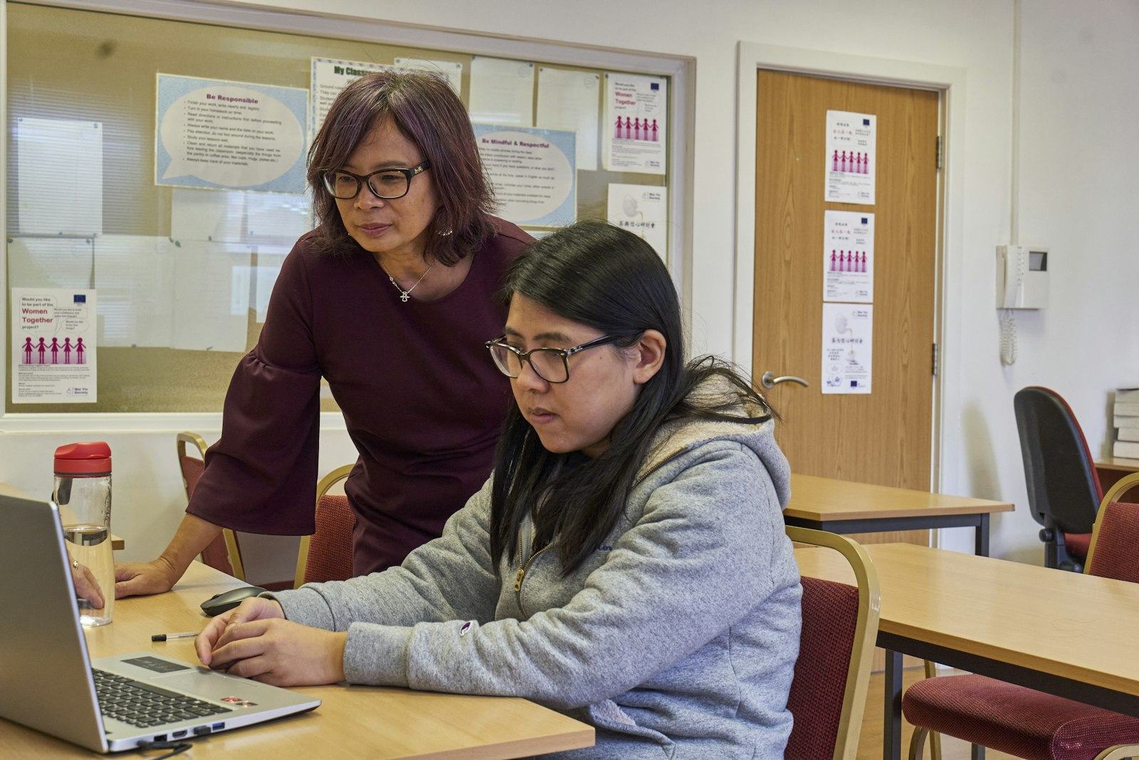 A teacher leans over to help a student working on a laptop in a classroom.