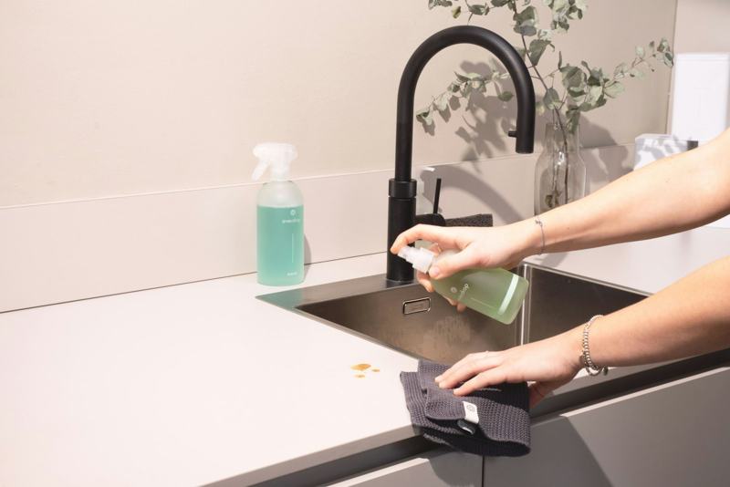 A hand spraying and wiping around a sink and black faucet