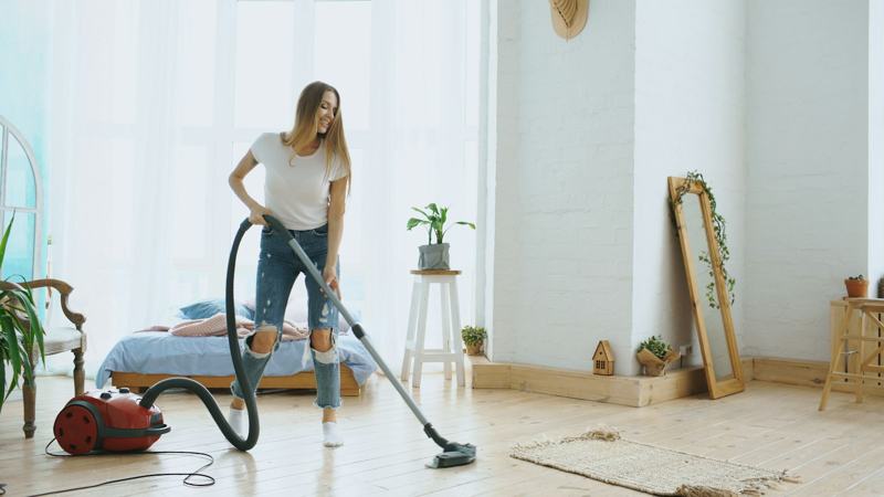 A woman vacuuming the wooden floor of a bright bedroom