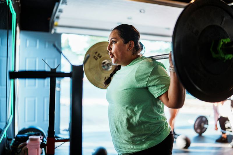 A woman holds a loaded barbell across her shoulders mid-squat in a bright gym