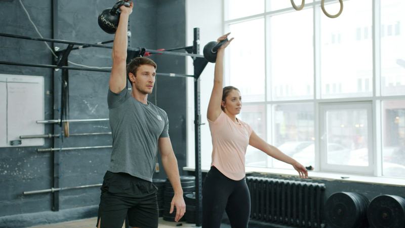 Two people finish a kettlebell swing with the bells overhead